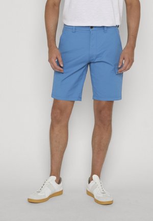 Homme portant un short décontracté bleu avec des poches latérales, des baskets blanches à semelles en gomme et un t-shirt blanc, debout devant un fond uni.