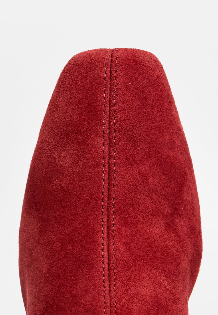 Czerwone zamszowe buty z wąskim czubkiem, gładką teksturą i wyraźnym przeszyciem wzdłuż środka, prezentujące elegancki i opływowy design.