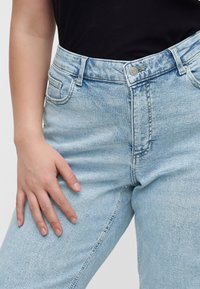 Personne portant un jean taille haute bleu clair et un haut noir, la main gauche reposant sur la cuisse.