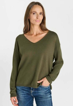 Donna con lunghi capelli castani che indossa un maglione verde oliva a scollo a V e jeans blu, in piedi con una mano in tasca davanti a uno sfondo bianco.