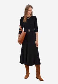 Czarna dopasowana sukienka z rękawami do trzech czwartek i paskiem w talii. Nosi ją w połączeniu z beżowymi zamszowymi botkami sięgającymi do kolan oraz trzymając brązową skórzaną torbę.