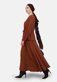 Brown, long-sleeve dress with a tie waist, flowing silhouette, and textured fabric. Paired with black stiletto heels and a dark patterned scarf.