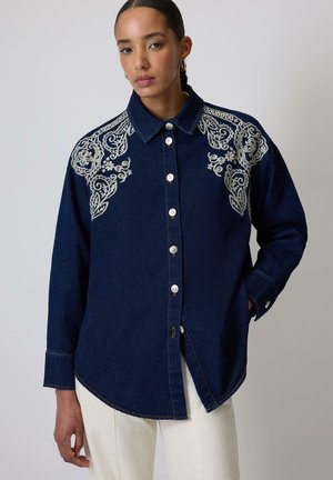 Navy denim shirt featuring white floral embroidery on the shoulders, button closure, and a curved hem. Long sleeves with button cuffs.