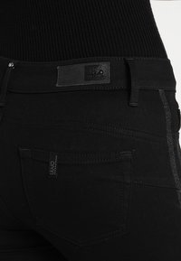 Svarta denimjeans med hög midja, läderlapp, subtila metalliska detaljer längs sidorna och bakfickor med varumärkeslogotyp.