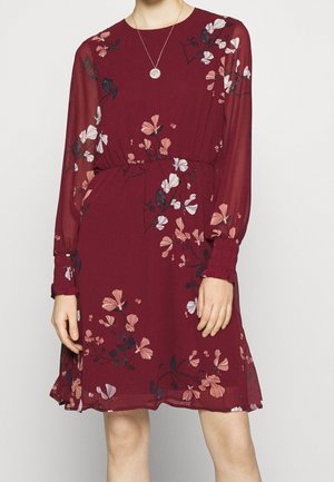 Kobieta ubrana w bordową sukienkę do kolan z długimi, przezroczystymi rękawami oraz różowo-białym kwiatowym wzorem, do tego założony okrągły naszyjnik z zawieszką.