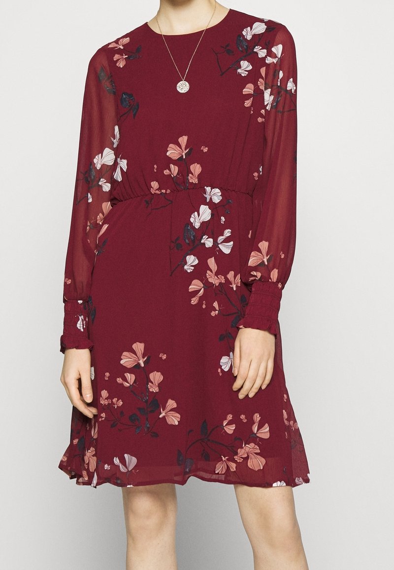 Femme portant une robe bordeaux longueur genoux avec des manches longues en tissu transparent et des motifs floraux roses et blancs, accessoirisée d'un collier pendentif rond.