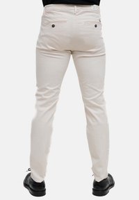 Pantaloni in cotone beige con un design slim fit, dotati di due tasche posteriori con bordi arrotondati e bottoni neri. Indossati con scarpe nere.