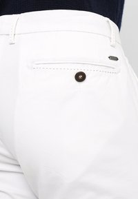 Witte broek met een achterzak met een bruine knop en subtiele stikdetails, gemaakt van een gladde, lichte stof.