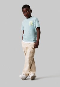 Camiseta de algodón azul claro con el logo amarillo "CK" combinada con pantalones cargo beige. Zapatillas blancas completan el conjunto, enfatizando un estilo informal.