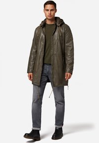 Olivgrön läderjacka med huva, med en dragkedjeförsedd bröstficka och snoddar. Matchas med grå jeans och svarta stövlar.