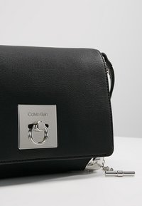 Sort lædertaske med en rektangulær sølvdetalje, der har en ring og kæde som pynt. Glat tekstur med en struktureret form.