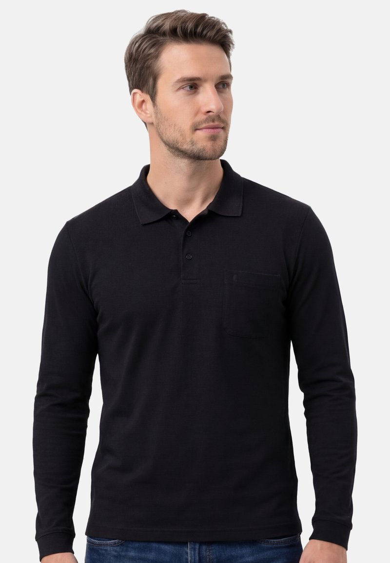 Homme portant un polo noir à manches longues avec une poche sur la poitrine, debout contre un fond clair uni, regardant sur le côté.