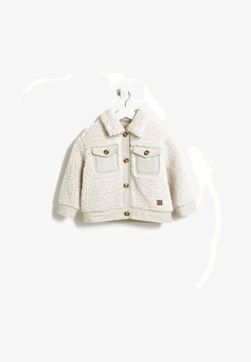 Veste en fausse fourrure crème au toucher doux, avec un col, deux poches avant, des boutons dorés et un écusson cousu en bas.