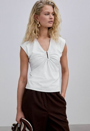 Femme blonde portant un haut blanc froncé sans manches, pantalon sombre, boucles d'oreilles créoles dorées, tenant un sac rayé sur un fond uni.