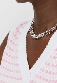 Witte gebreide trui met een roze geruit patroon en een v-halslijn. Zilveren ketting voegt textuur toe. Close-up weergave.