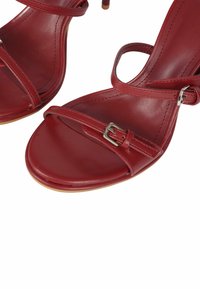 Sandali rossi in pelle con un design a cinghie sottili, punta aperta e tacco piccolo. Presentano dettagli decorativi con fibbie sulle cinghie alla caviglia.