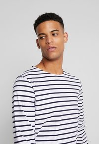 T-shirt a maniche lunghe bianco e blu a righe, con collo a giro e una texture di tessuto morbida. Vestibilità casual con cuciture delle spalle visibili.