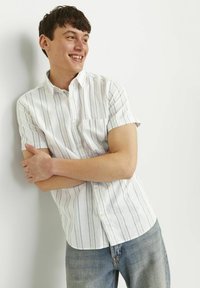 Camisa de manga corta abotonada en blanco con rayas verticales azules, con cuello y un bolsillo en el pecho. Llevada con jeans de mezclilla azul claro.