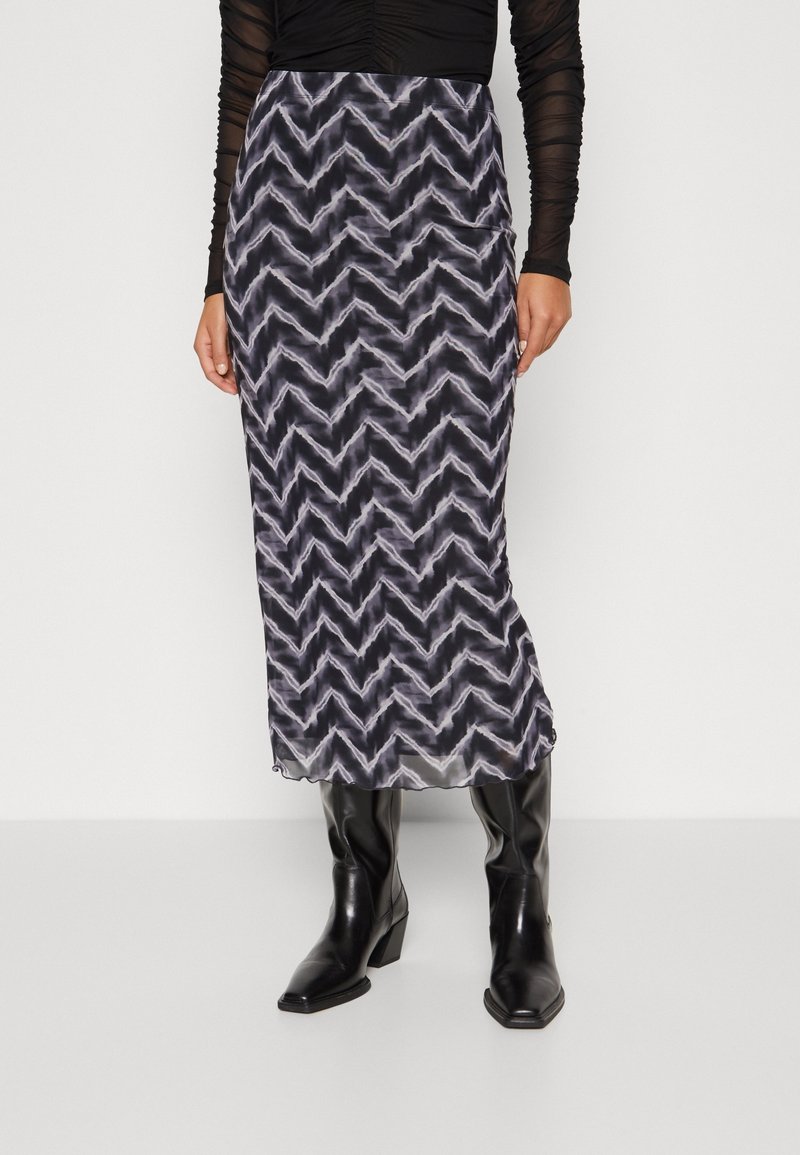 Femme portant une jupe midi à motif zigzag noir et blanc avec texture transparente, un top froncé noir et des bottes en cuir noires.
