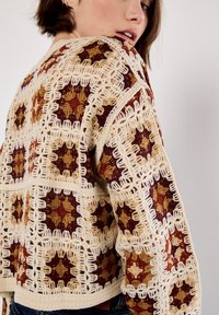 Knit cardigan featuring a beige base with intricate geometric patterns in gold, rust, and brown, accented with textured crochet trim.