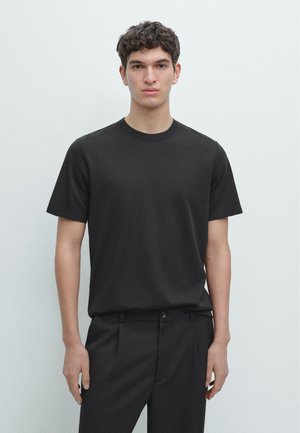 Ung mand står op ad en hvid væg iført en sort kortærmet rundhals t-shirt og sorte bukser, vendt fremad.