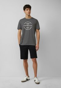 Grijze katoenen t-shirt met witte ronde logoprint, gecombineerd met zwarte shorts en witte sneakers met zwarte accenten. Eenvoudig, casual ontwerp.