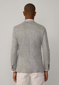 Blazer grigio con texture, dal taglio sartoriale, con revers a punta, due bottoni e dettagli cuciti. Indossato sopra una camicia chiara e pantaloni crema.