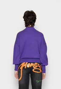 Maglione a maglia viola con colletto alto e spalle ribassate, abbinato a jeans neri con testo grafico arancione e verde sul retro.