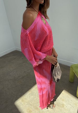 Γυναίκα που φοράει φόρεμα ροζ και κόκκινο με μοτίβο χωρίς ώμους, κρατώντας ένα τσαντάκι με μαύρο και λευκό μοτίβο, στέκεται σε σκυρόδετο δάπεδο.