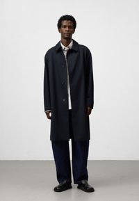 Cappotto trench nero, abbottonato, con colletto, indossato sopra una camicia a righe e pantaloni in denim scuri e larghi. Tessuto liscio, design fino al ginocchio.