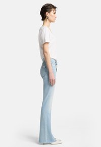 Jeans svasati di un azzurro chiaro presentano un design sbiadito, vita alta e orlo grezzo, abbinati a una maglietta bianca aderente e scarpe bianche.