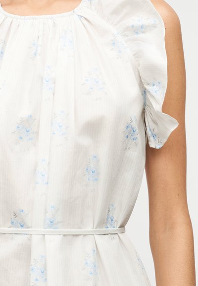 Λευκό αμάνικο φόρεμα με ανοιχτό γαλάζια floral σχέδια, βολάν στο ύψος των ώμων και λεπτή ζώνη στη μέση, σε ένα άτομο με μαυρισμένο δέρμα.