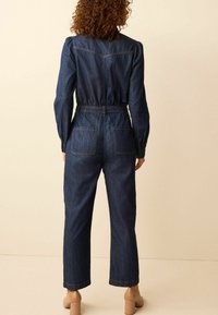 Combinaison en denim avec une taille cintrée, des manches longues et des poches arrière. Présente une couleur bleu foncé et des détails de couture visibles.