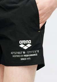 Μαύρο σορτς με λευκό λογότυπο «arena» και συντεταγμένες, φορεμένο από ένα άτομο με το χέρι μέσα στην δεξιά τσέπη.