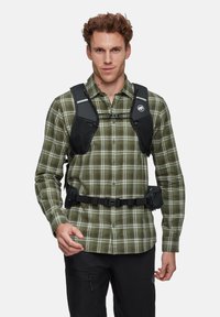 Imbracatura nera con cinture regolabili sopra una camicia verde a scacchi. Presenta imbottiture in rete e una tasca laterale. Un uomo sta in piedi con sicurezza.