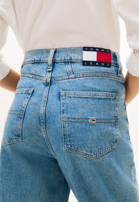 Zbliżenie na niebieskie dżinsy Tommy Jeans z tylnymi kieszeniami i naszywką z logo, noszone z białą włożoną koszulą.