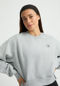 Sudadera gris con cuello redondo, puños y dobladillo acanalados. Presenta un pequeño logotipo circular en el pecho. Textura suave y corte relajado.