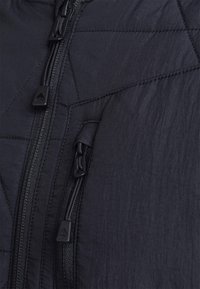 Svart isolerad jacka med kviltat mönster, framdragkedjor och strukturerad tyg. Designen inkluderar subtila snedställda sömmar och mattsvart metall.