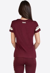 Camiseta de manga corta color marrón, hecha de algodón, con rayas blancas en las mangas y un pequeño logo en la parte posterior del cuello. Textura suave.