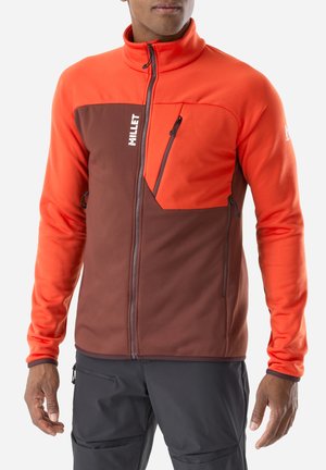 Man wearing a brown and bright orange zip-up fleece jacket with chest and side zip pockets, standing against a plain background.