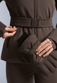 Hnedý vonkajší odev s textúrovaným pásom, zipsovými vreckami a zateplenými rukavicami. Obsahuje klip na bezpečné pripnutie doplnkov.