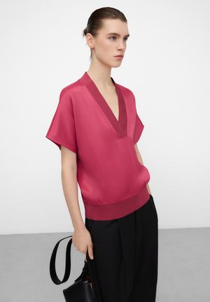 Kobieta z zaczesanymi do tyłu włosami, ubrana w satynową bluzkę w kolorze fuksji z głębokim dekoltem w serek oraz czarne spodnie, trzymająca czarną torebkę obok siebie.