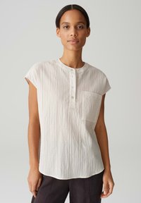 Camicia corta a maniche corte beige chiaro con scollatura rotonda, tessuto a trama, taschino frontale e tre bottoni sulla patta.