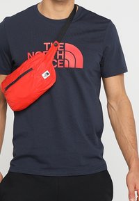 Sac bandoulière en nylon rouge avec une sangle noire ; il présente un compartiment zippé et un accent logo, porté par-dessus un T-shirt bleu marine avec un logo bien visible.