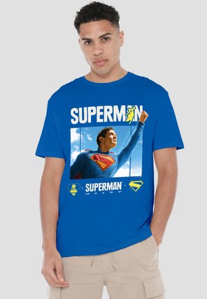Young man wearing blue Superman t-shirt with Superman flying graphic and beige shorts against plain background.