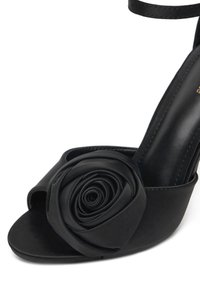 Czarna sandałka na wysokim obcasie z dużą tkaninową różą na przodzie, gładkim satynowym wykończeniem, otwartym noskiem i paskiem wokół kostki.