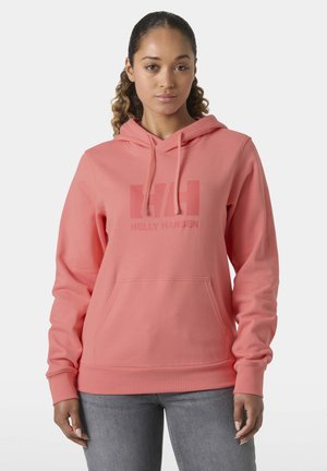 Mujer con cabello rizado que lleva una sudadera con capucha coral de Helly Hansen y jeans grises, de pie contra un fondo claro y liso.
