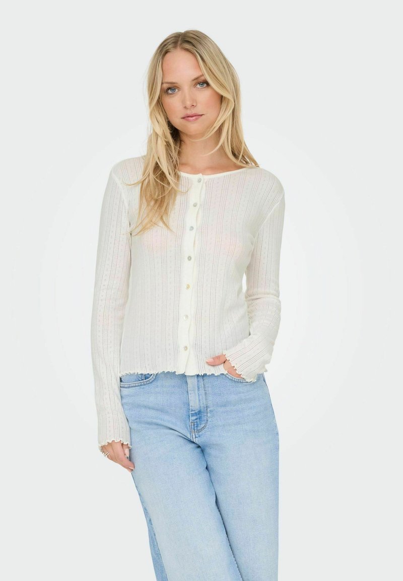 Witte geribde cardigan met knoopsluiting, gekartelde manchetten en zoom. Draag het over lichtblauwe jeans met een textuurpatroon.