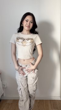 Νεαρή γυναίκα με μακριά σκούρα μαλλιά, φοράει μπεζ crop top και παντελόνια cargo, στέκεται μπροστά από μονόχρωμο τοίχο, με τα χέρια κοντά στους γοφούς, χαμογελώντας ελαφρώς.