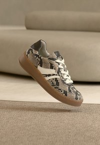 Αθλητικό παπούτσι με υφή σε μαύρο και μπεζ σχέδιο φιδιού. Διαθέτει λευκή λωρίδα, μαύρη ταμπέλα στη φτέρνα και ελαφρώς καφέ λαστιχένια σόλα.